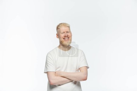 bärtiger Albino-Mann im T-Shirt, lächelnd und mit verschränkten Armen auf weißem Grund stehend 