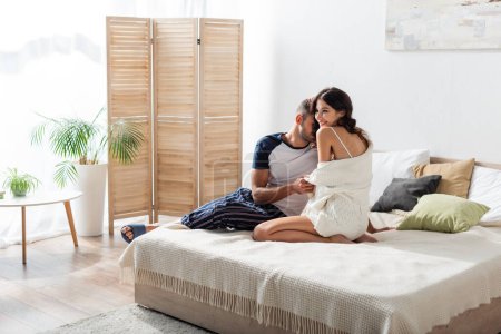 Téléchargez les photos : Bearded man kissing smiling girlfriend in cardigan on bed at home - en image libre de droit
