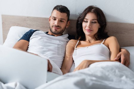 Téléchargez les photos : Man hugging girlfriend and looking at blurred laptop on bed - en image libre de droit