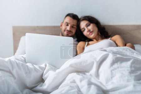 Téléchargez les photos : Laptop near blurred couple lying on bed in morning - en image libre de droit