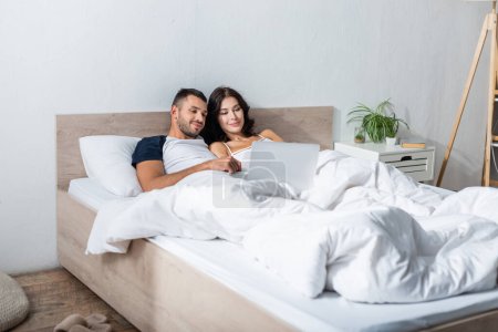 Téléchargez les photos : Brunette couple looking at laptop while lying on bed in morning - en image libre de droit