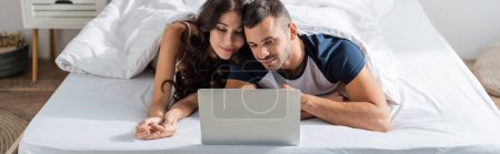Téléchargez les photos : Brunette couple looking at laptop on bed at home, banner - en image libre de droit