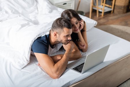 Téléchargez les photos : Brunette couple looking at laptop on bed while relaxing at home - en image libre de droit