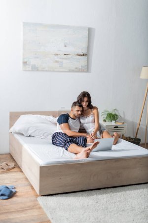 Téléchargez les photos : Couple in pajama holding hands near laptop on bed in morning - en image libre de droit