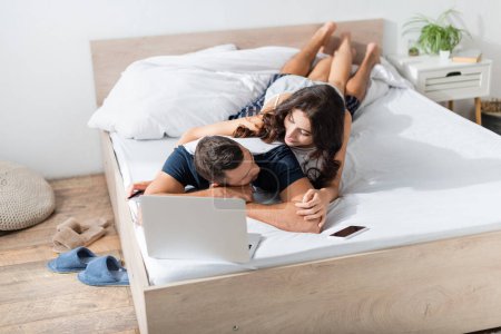 Brunette woman hugging boyfriend near devices on bed at home 