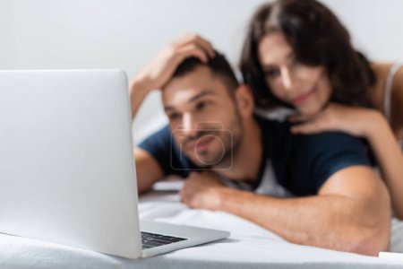 Téléchargez les photos : Laptop near blurred couple lying on bed at home - en image libre de droit