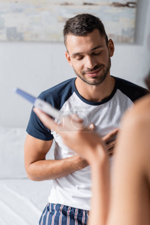 Smiling man in pajama standing near girlfriend with pregnancy test in bedroom 