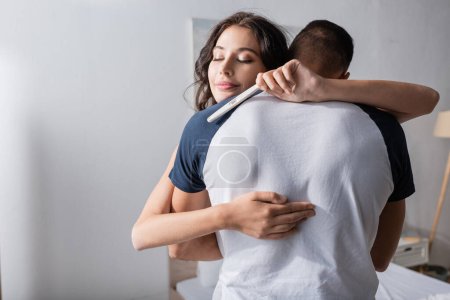 Smiling woman holding pregnancy test and hugging boyfriend at home 