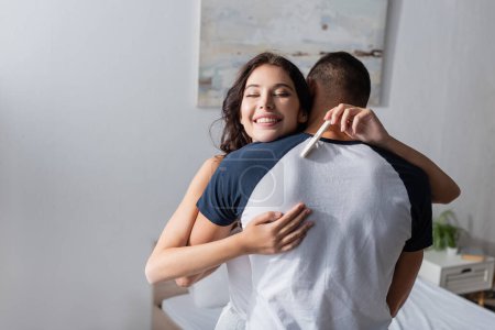 Happy woman holding pregnancy test and embracing boyfriend in bedroom 