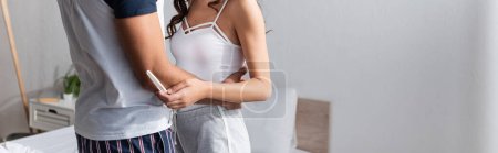 Cropped view of man in pajama hugging girlfriend with pregnancy test at home, banner 