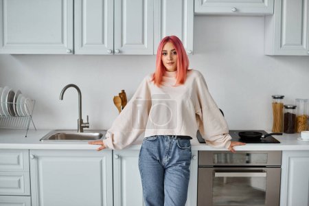 Foto de Mujer alegre de buen aspecto en jersey casual y jeans posando en la cocina y mirando a la cámara - Imagen libre de derechos