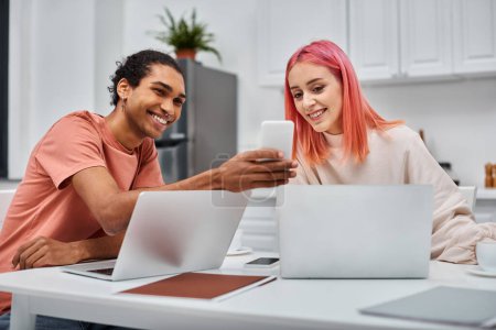 Photo for Happy loving multiracial couple in casual attires working at laptops and looking at smartphone - Royalty Free Image