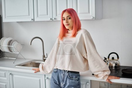 cheerful attractive woman in white casual jumper and jeans looking at camera while in kitchen