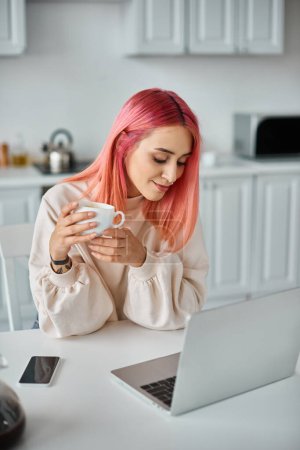 Foto de Atractiva mujer elegante con pelo rosa trabajando en el ordenador portátil bebiendo café caliente y mirando hacia otro lado - Imagen libre de derechos