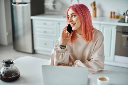 Téléchargez les photos : Gai belle femme avec les cheveux roses en tenue décontractée parler par téléphone tout en travaillant avec ordinateur portable - en image libre de droit