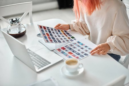 Photo for Cropped view of young woman with pink hair holding papers with images next to her laptop at home - Royalty Free Image