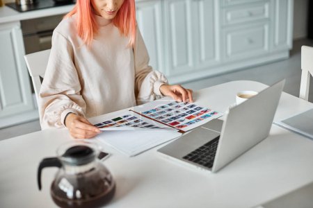 cropped view of woman in casual attire holding papers with images next to her laptop and coffee