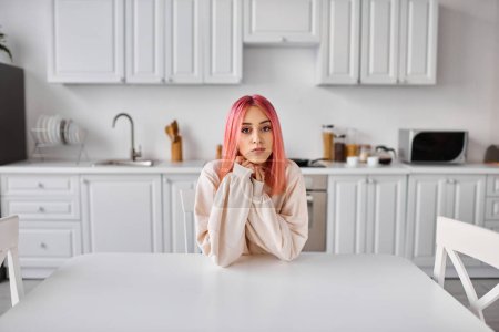 Foto de Hermosa mujer joven con el pelo rosa sentado en la cocina en la mesa y mirando directamente a la cámara - Imagen libre de derechos