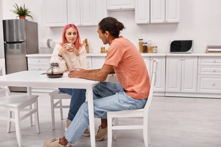 Foto de Atractiva pareja multirracial alegre en ropa de casa sentado en la cocina y sonriendo el uno al otro - Imagen libre de derechos