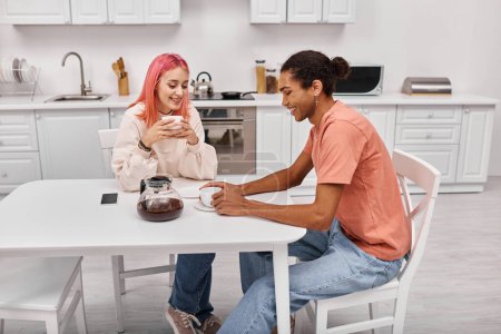 Photo for Good looking cheerful diverse couple sitting at table with coffee and looking at each other - Royalty Free Image