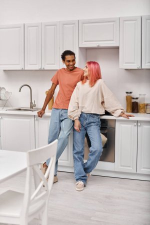 Foto de Atractiva pareja multirracial alegre en ropa de casa sonriendo felizmente el uno al otro en la cocina en casa - Imagen libre de derechos