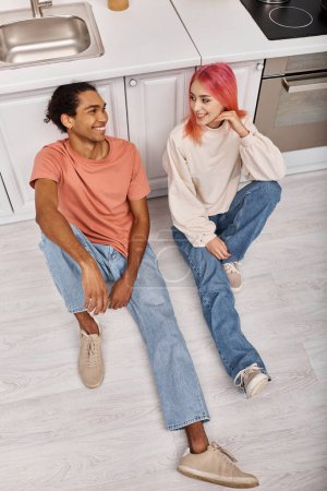 Photo for Beautiful multicultural joyous couple in casual attires sitting on floor and smiling at each other - Royalty Free Image
