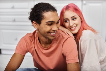 Foto de Enfoque en el hombre americano africano alegre en ropa de casa sonriendo a su novia de pelo rosa borrosa - Imagen libre de derechos