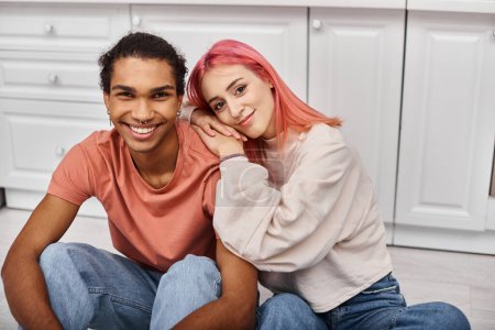 Téléchargez les photos : Joyeux couple multiculturel attrayant dans des vêtements confortables souriant joyeusement à la caméra dans la cuisine à la maison - en image libre de droit