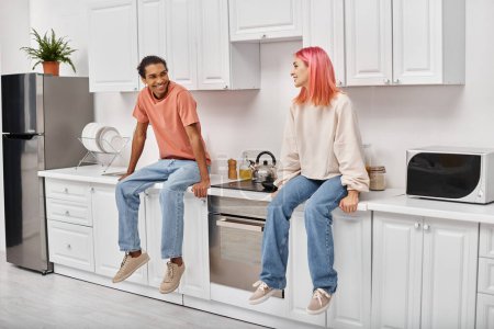 Foto de Alegre atractiva pareja multirracial en ropa de casa sentado en el mostrador de la cocina y sonriendo el uno al otro - Imagen libre de derechos