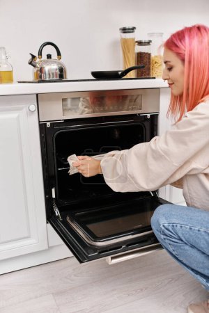 junge attraktive Frau mit rosa Haaren in lässiger, gemütlicher Kleidung mit Backofen, um zu Hause das Abendessen zu kochen
