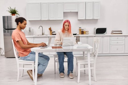 appealing hard working interracial couple sitting at table in kitchen with laptops, freelance