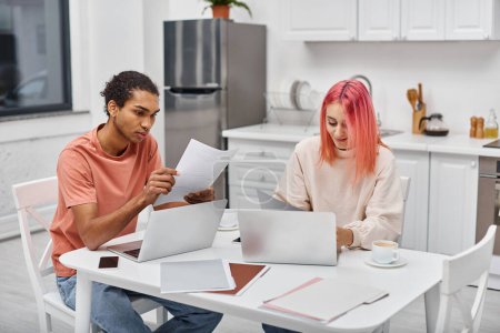 gut aussehendes multiethnisches Paar in Homewear, das zu Hause hart mit Papierkram und Laptops arbeitet