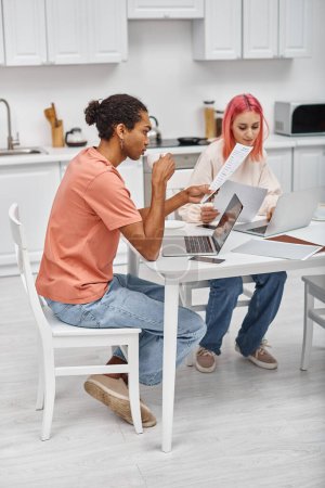 Foto de Atractivo joven pareja multicultural sentado en las computadoras portátiles y trabajando con el papeleo en casa - Imagen libre de derechos
