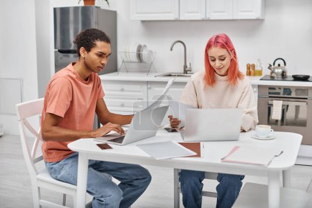 Photo for Beautiful young interracial couple in homewear working with paperwork and laptops in kitchen at home - Royalty Free Image