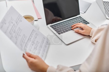 Foto de Vista recortada de mujer joven en traje casual sosteniendo su papeleo mientras trabajaba en el ordenador portátil en casa - Imagen libre de derechos