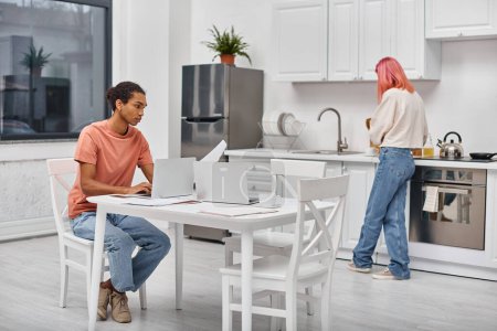 Téléchargez les photos : Bel homme afro-américain en vêtements de maison travaillant à l'ordinateur portable à côté de sa petite amie aux cheveux roses - en image libre de droit