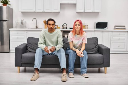 Téléchargez les photos : Joyeux couple multiracial en tenue décontractée assis sur le canapé et regardant la caméra à la maison - en image libre de droit