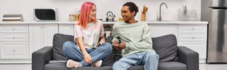 Photo for Appealing multicultural jolly couple in homewear sitting on sofa and smiling at each other, banner - Royalty Free Image