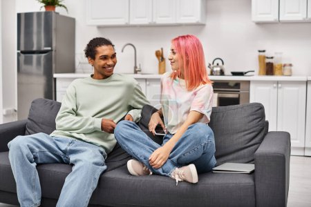 Foto de Atractiva pareja interracial alegre en ropa de casa sentado en el sofá y sonriendo felizmente el uno al otro - Imagen libre de derechos