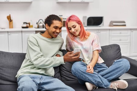Foto de Alegre pareja multirracial en ropa casual sentado en el sofá y mirando el teléfono inteligente en casa - Imagen libre de derechos