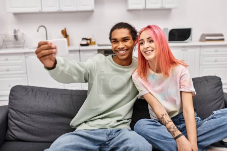 Foto de Alegre pareja multicultural atractiva en la ropa de casa tomando selfies mientras que en la sala de estar en casa - Imagen libre de derechos