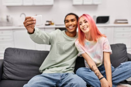 Foto de Centrarse en el teléfono inteligente en las manos en la alegre pareja diversa tomando selfies mientras está en la sala de estar en casa - Imagen libre de derechos
