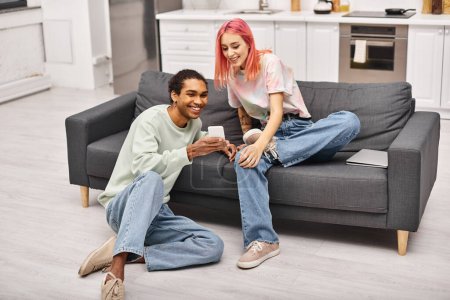Foto de Alegre atractiva pareja interracial en ropa de casa sentado en la sala de estar y mirando el teléfono inteligente - Imagen libre de derechos