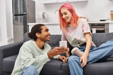 Photo for Beautiful joyful multiracial couple in homewear sitting in living room and looking at phone happily - Royalty Free Image