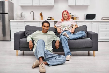 Foto de Alegre atractiva pareja multirracial en acogedora ropa de casa posando en la sala de estar y sonriendo a la cámara - Imagen libre de derechos