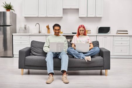 Foto de Alegre pareja interracial atractiva en la ropa de casa sentado en el sofá y trabajando en computadoras portátiles de forma remota - Imagen libre de derechos