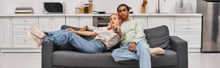 Foto de Atractiva pareja interracial en ropa de casa pasar tiempo juntos y ver películas en casa, pancarta - Imagen libre de derechos