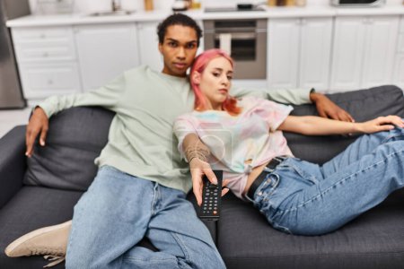 focus on remote control in hand of pretty blurred woman and her african american boyfriend at home