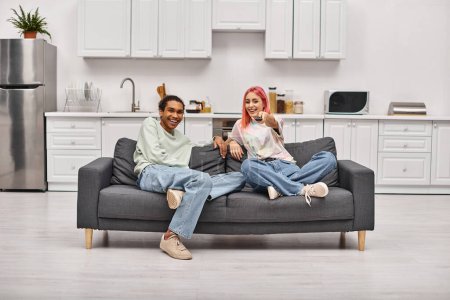 Téléchargez les photos : Joyeux beau couple diversifié en vêtements décontractés assis sur le canapé et regarder des films ensemble - en image libre de droit