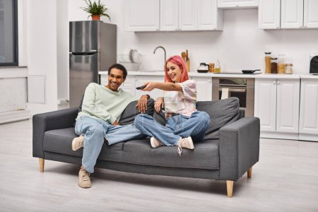 Foto de Alegre atractiva pareja diversa en trajes acogedores sentados en el sofá y ver películas juntos - Imagen libre de derechos
